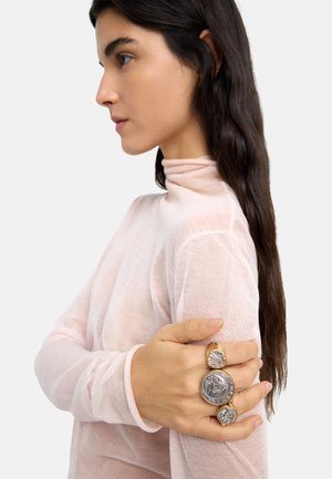 Kobieta z długimi ciemnymi włosami, ubrana w przezroczysty, bladoróżowy golf, trzymająca się za ramię i prezentująca efektowny pierścionek z trzema dużymi, okrągłymi monetami.