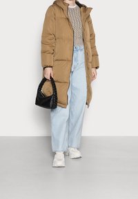 Giacca puffer marrone con cappuccio, dotata di zip frontale. Indossata sopra un maglione beige testurizzato e jeans a gamba larga azzurri. Borsa nera intrecciata.