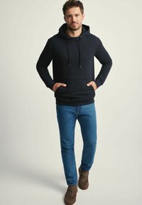 Mężczyzna w czarnej bluzie z kapturem, niebieskich dżinsach i brązowych trampkach stoi z rękami w kieszeni bluzy na jasnym tle.