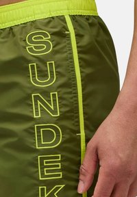 Shorts verts en matière légère avec des accents jaunes, portant le mot « SUNDEK » brodé en grandes lettres sur le côté.