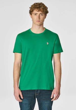 T-shirt verde in cotone con collo rotondo e maniche corte; presenta un piccolo logo rosa sul lato sinistro del petto. Indossato con jeans blu.