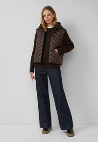 Donna che indossa un gilet imbottito marrone scuro sopra un maglione marrone, jeans a gamba larga scuri e stivali neri con suole beige, in piedi contro un muro neutro.