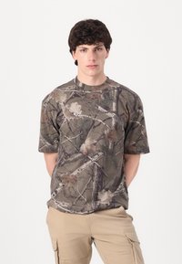 T-shirt camouflage dans des tons terreux avec un motif de feuilles et de branches. Manches courtes et col rond. Fabriqué à partir d'un tissu léger. Coupe décontractée.