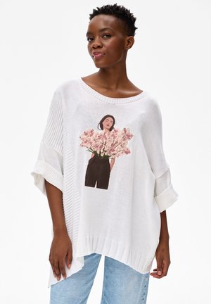 Kobieta w luźnym białym swetrze z ilustracją osoby trzymającej różowe kwiaty, w jasnoniebieskich dżinsach, stoi na białym tle.