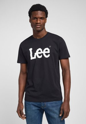 Homme portant un t-shirt noir avec le logo Lee et un jean bleu, debout devant un fond gris uni.