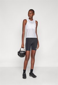 Hvit ermeløs treningsskjorte med mesh-design, kombinert med svarte sykkelshorts. Modellen holder en svart sykkelhjelm og har på seg sykkelsko.