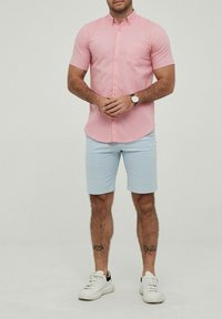 Chemise boutonnière à manches courtes de couleur rose clair avec une poche sur la poitrine, assortie à un short bleu clair. Des baskets blanches complètent la tenue.