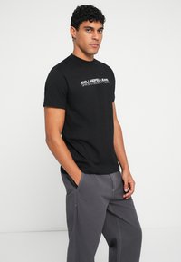 Melna kokvilnas T-kreklu ar apaļu kaklu un īsām piedurknēm; priekšpuses dizainā ir atspoguļots "KARL LAGERFELD JEANS" kontrastējošā baltā tekstā.