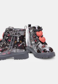 Botine negre din piele patentată, cu text imprimat și inimioare. Au tălpi din cauciuc gri, șireturi negre și un fermoar pe lateral pentru a fi ușor de purtat.