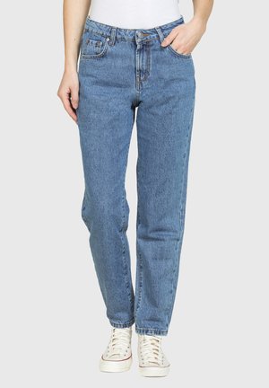 Persona che indossa jeans blu a vita alta con gambe dritte, con la mano in tasca e sneaker bianche, in piedi davanti a uno sfondo chiaro e uniforme.