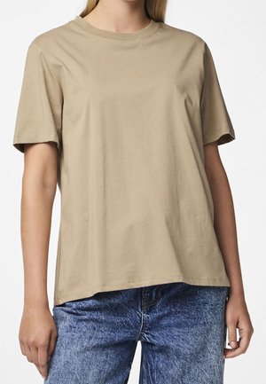 Femme portant un t-shirt beige uni à manches courtes et un jean bleu délavé à l'acide, debout sur un fond blanc.