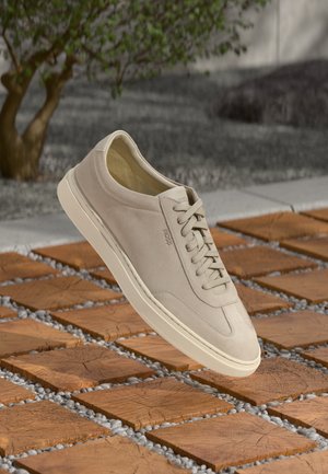 Sneaker in suede beige con suola e lacci crema, con la scritta "BOSS" in rilievo, fluttua sopra un pavimento esterno in legno e ciottoli.