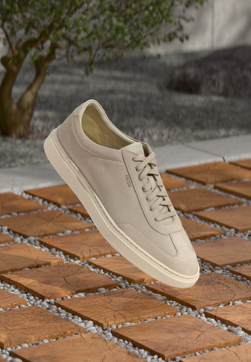 Beige suede sneaker with cream sole and laces, embossed "BOSS," floating above wooden block and pebble outdoor flooring.