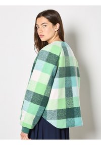 Jeune femme portant une veste en polaire à carreaux verts et bleus, regardant par-dessus son épaule sur un fond blanc uni.