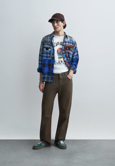 Καρό πουκάμισο σε μπλε και καφέ χρώματα με καλλιτεχνικά σχέδια προσώπου πάνω από ένα λευκό γραφιστικό μπλουζάκι. Καφέ παντελόνι και αθλητικά παπούτσια με πετρόλ λεπτομέρειες. Καφέ καπέλο.