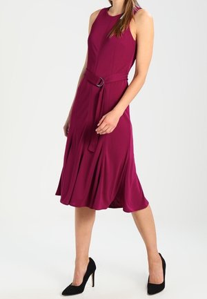 Vrouw draagt mouwloze knielange magenta jurk met riem en zwarte hoge hakken, staand voor een effen achtergrond.