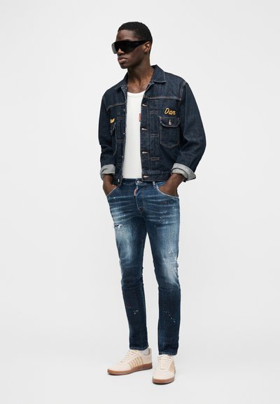 Donker denim jack met gele borduurwerk, wit tanktopje, versleten blauwe spijkerbroek en beige sneakers met bruine accenten. Grote zwarte zonnebril.
