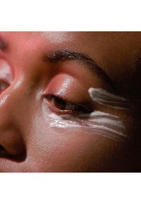 Close-up of an eye with a smooth, white cream applied underneath. Skin tone is deep brown, with highlighted areas and a soft, glossy texture.