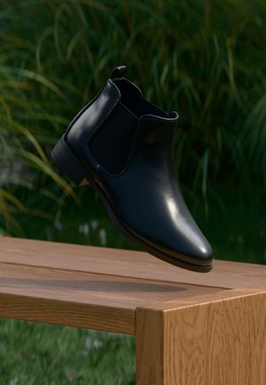 Zwarte leren chelseaboot met elastische zijkanten, een gladde afwerking, ronde neus en een lage gestapelde hak, weergegeven boven een houten oppervlak.