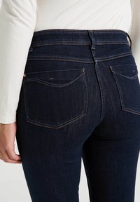 Jeans de mezclilla azul oscuro con una cintura de altura media, bolsillos traseros con costuras sutiles y una textura suave. Los acentos incluyen detalles de hardware en color cobre.