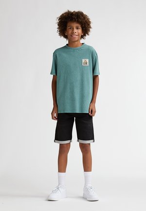 Türkisfarbenes Baumwoll-T-Shirt mit einem vorderen Grafik-Patch, kombiniert mit schwarzen Denim-Shorts und weißen Turnschuhen, vor einfarbigem Hintergrund.