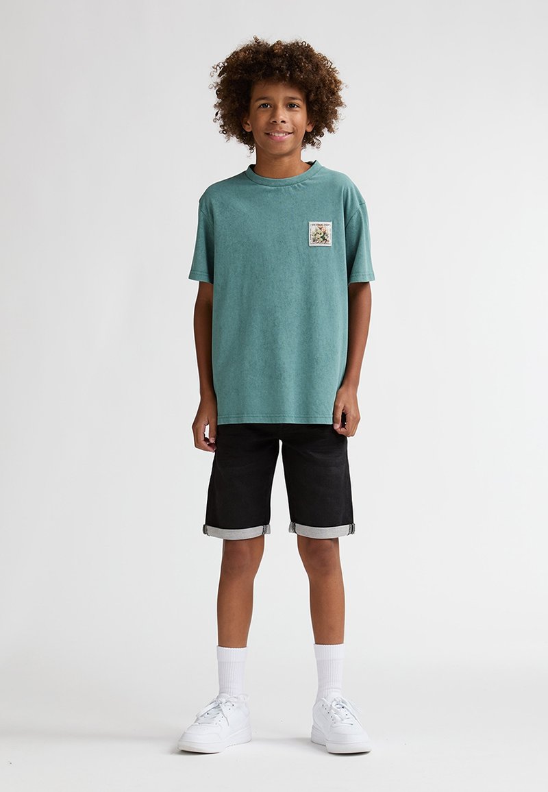 Tyrkis bomuld t-shirt med et grafisk patch foran, kombineret med sorte denimshorts og hvide sneakers, på en ensfarvet baggrund.