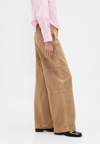 Pantalon à jambes larges en denim marron clair, avec un design en patchwork et des détails de coutures visibles. Associé à une chemise rayée rose.