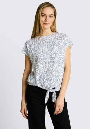 Kobieta o długich blond włosach, ubrana w białą bluzkę z krótkim rękawem z niebieskim kwiatowym wzorem i wiązaniem z przodu, połączoną z czarnymi spodniami.