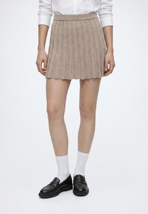 Femme portant une mini-jupe en maille côtelée beige, des chaussettes blanches, des mocassins noirs et une chemise blanche à manches longues sur un fond uni.