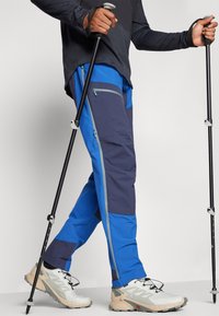 Blue and navy hiking pants with zippered pockets, paired with beige and gray athletic shoes. Featuring a textured fabric and adjustable straps.