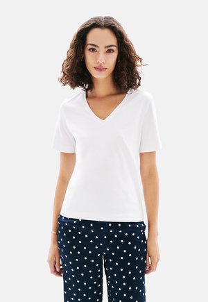 Weißes Kurzarm-V-Ausschnitt-T-Shirt aus glattem Stoff, getragen mit navyblauen Polka-Dot-Hosen, mit einem lässigen Schnitt und dezentem Stichdetail.