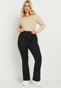 Mustat denim-housut leveällä lahkeella ja korkealla vyötäröllä, joissa on vyölenkit ja taskut, yhdistettynä beigeen pitkähiharaiseen paitaan. Valkoiset tennarit jalkineina.