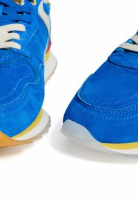 Niebieskie zamszowe sneakersy z białymi sznurówkami, żółtymi i czerwonymi akcentami oraz teksturowanym materiałem. Posiadają gładką podeszwę z przezroczystą krawędzią.