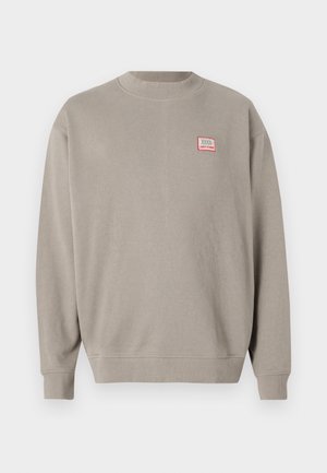 Sweat-shirt gris clair en tissu doux avec un col rond et des poignets côtelés. Il affiche un petit patch logo rouge et blanc sur la poitrine.