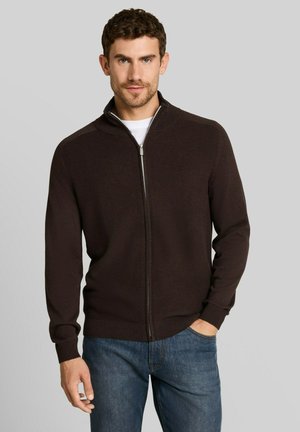 Mann mit kurzen lockigen Haaren trägt einen dunkelbraunen Reißverschluss-Pullover über einem weißen Hemd und blauen Jeans, steht mit einer Hand in der Tasche.