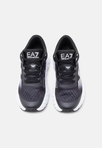 Fekete sportcipők texturált szintetikus felsőrésszel, fehér gumi talppal és logóval a nyelvén. Kontrasztos dizájnelemek jellemzik.