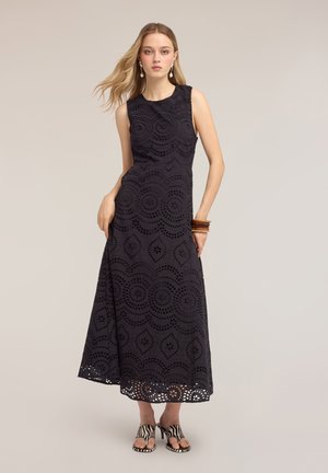 Femme portant une robe maxi en dentelle noire sans manches avec des motifs circulaires, des bracelets en bois, des boucles d'oreilles pendantes et des sandales à talons imprimé zèbre.