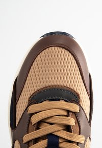 Brown and beige sneaker featuring textured fabric, smooth leather accents, and beige laces. Includes a navy detail near the tongue.