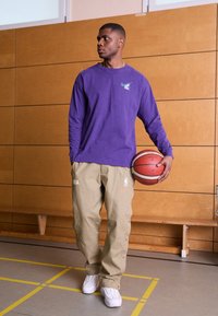 Una maglietta a maniche lunghe viola con logo verde, pantaloni khaki con accenti NBA e Nike, che tiene in mano un pallone da basket, in piedi su un pavimento da palestra.
