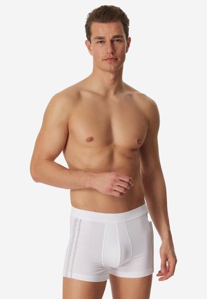Gespoorde man met kort haar, gekleed in witte boxershorts met dubbele grijze strepen aan de zijkanten, staand tegen een effen achtergrond.