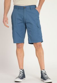 Pantalones cortos de carga azules con cierre de botón, bolsillos laterales y corte recto. Combinados con zapatillas altas negras y una camiseta gris.