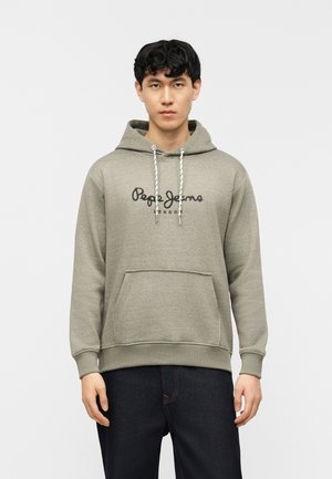 Mladi moški, ki nosi sivo kapucar s sprednjim žepom blagovne znamke Pepe Jeans London in temne kavbojke, stoji pred enostavnim svetlim ozadjem.