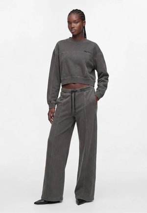 Vrouw met gevlochten haar, gekleed in een bijpassende donkergrijze cropped trui en wijde broek met zwarte puntige schoenen, staand met één hand in de zak.