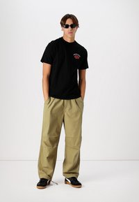 Black cotton t-shirt with red graphic print, paired with khaki wide-leg trousers, featuring pockets and elastic cuffs, worn with black sneakers.
