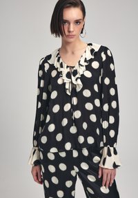 Blusa nera con pois bianchi, scollatura arricciata, dettaglio con fiocco e maniche a campana. Realizzata in tessuto morbido con una vestibilità fluida.
