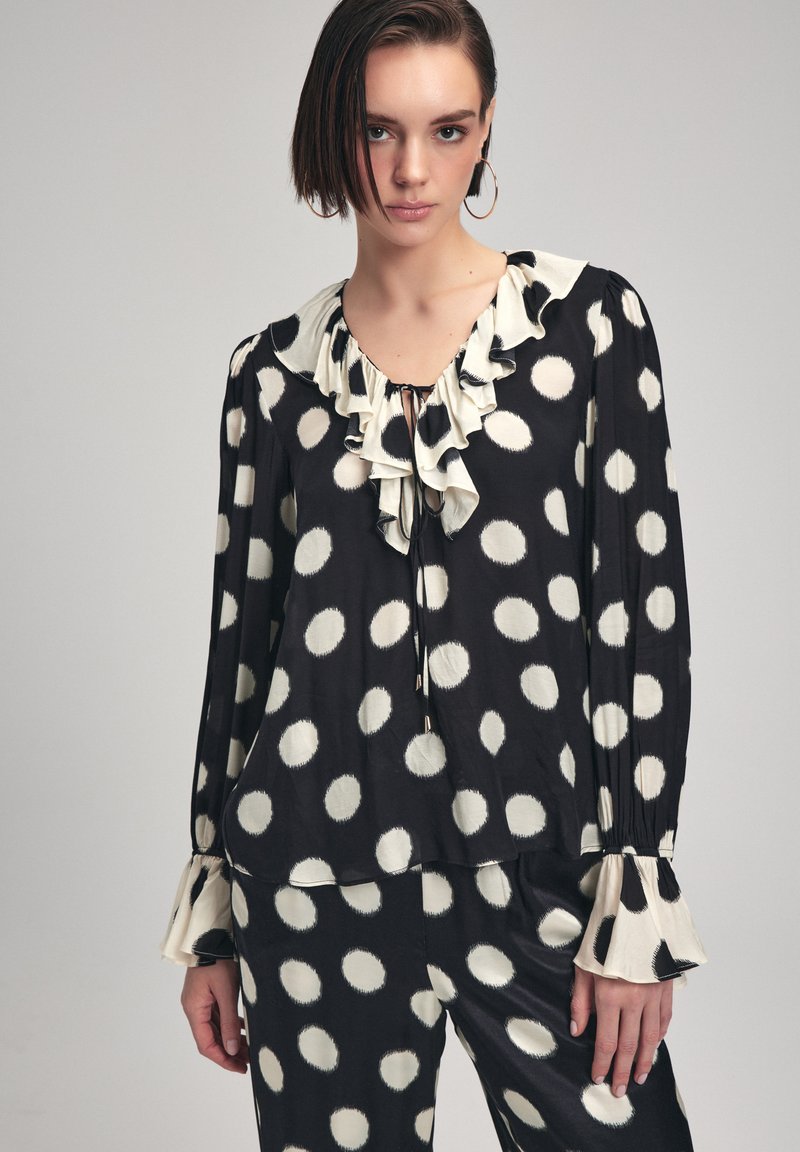Blusa nera con pois bianchi, scollatura arricciata, dettaglio con fiocco e maniche a campana. Realizzata in tessuto morbido con una vestibilità fluida.