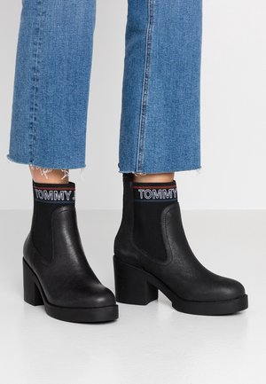 Botas negras hasta el tobillo con un tacón grueso, hechas de cuero texturizado, que cuentan con paneles laterales elásticos y una banda con el logo en la parte superior.