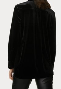 Czarna koszula z aksamitu z długimi rękawami o luźnym kroju, z zakładką na plecach i mankietami na guziki. Gładka tekstura z minimalnie widocznymi szwami.