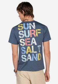 Camiseta de algodón azul con un gran gráfico de texto multicolor: "SOL OLA SAL MAR Arena". Manga corta y un diseño clásico de cuello redondo.