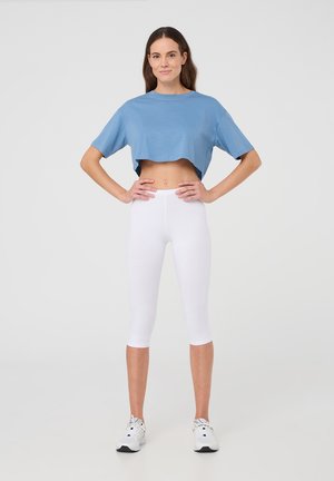 T-shirt in cotone blu tagliato, con maniche corte, abbinato a leggings bianchi fino al polpaccio. Il modello indossa scarpe da ginnastica bianche. Sfondo neutro.
